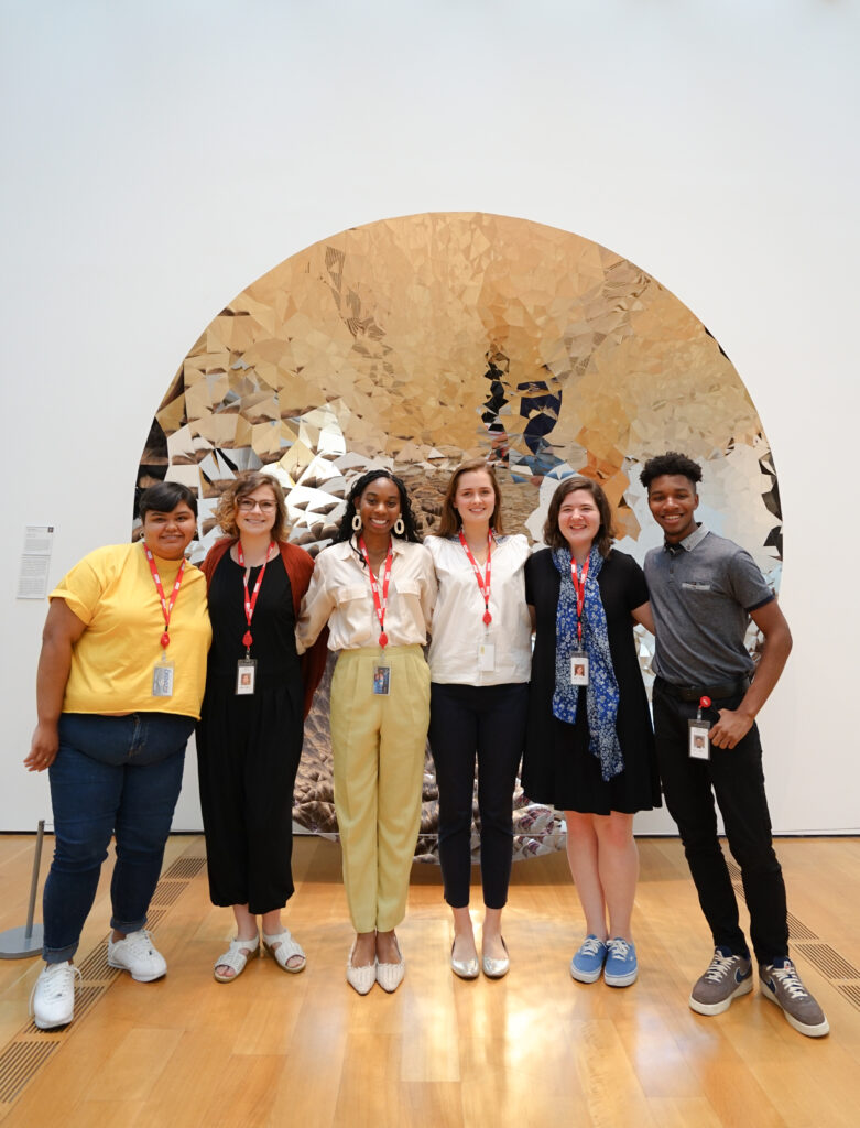 Six High Museum Summer Interns stand in front of a mirrored Anish Kapoor artwork.