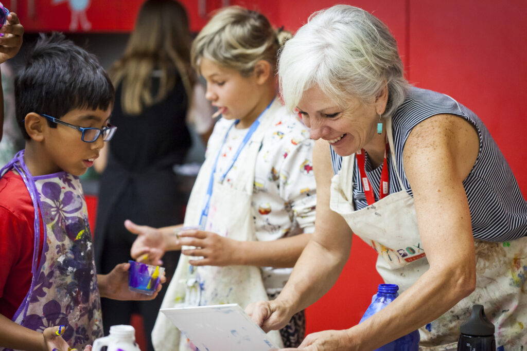 An educator in an apron shows a student how to finger paint at Summer Art Camp