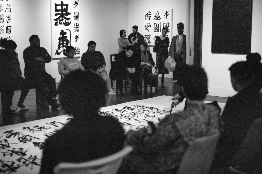 Brandon Sadler Banner Image A group of people watch an artist create large calligraphy on paper in a gallery filled with bold Chinese calligraphy art on the walls; the audience sits and stands, observing intently.