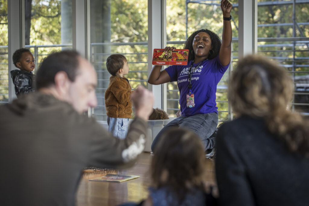 Toddler Storytime Family Saturday First Saturday A woman kneels on the floor, energetically holding up a colorful game box to a small group of children and adults in a bright room with large windows. Some children and adults watch her with interest.