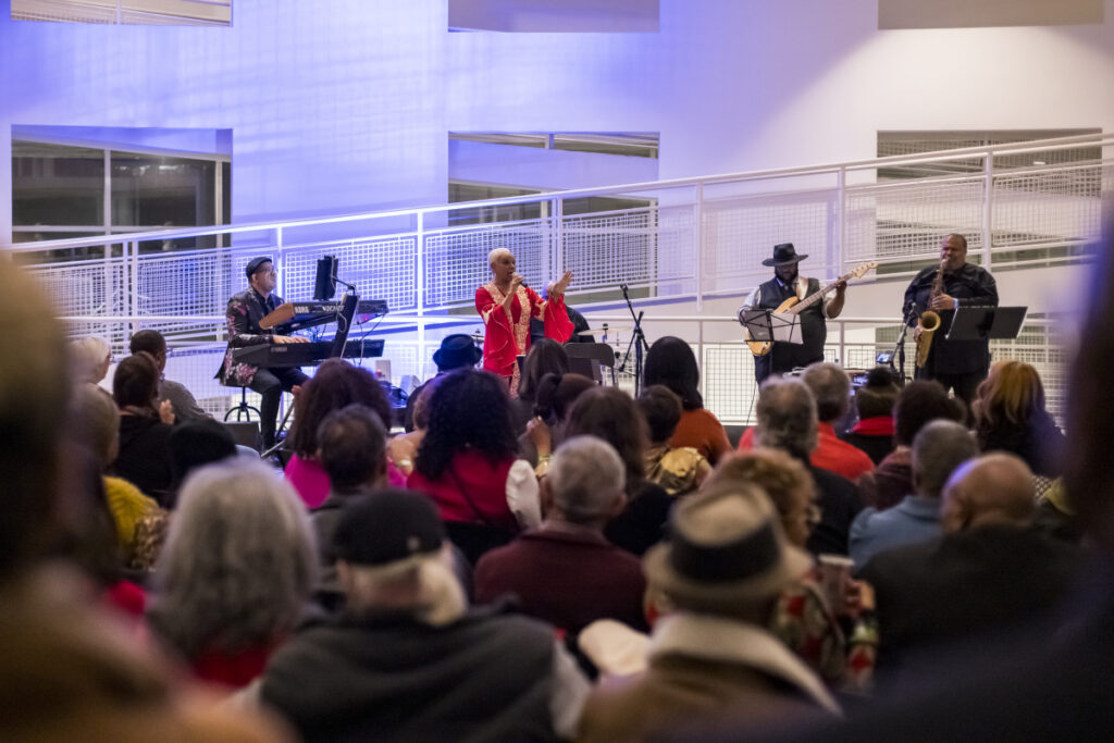 High Friday Jazz Gd1a0788 O3 A jazz band performs on stage for a seated audience in a modern indoor venue, with the singer in red and musicians playing keyboard, guitar, and drums. The audience watches attentively.