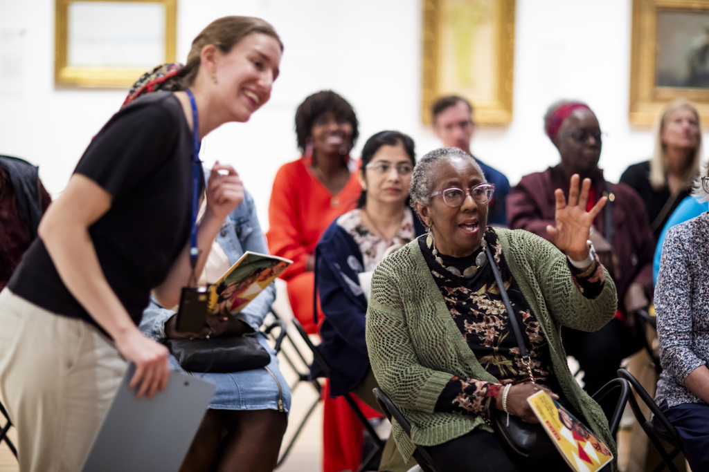 A group of people sit and listen attentively in an art gallery; one woman in a green sweater gestures while speaking, as another woman beside her smiles and observes. Paintings hang on the wall in the background.