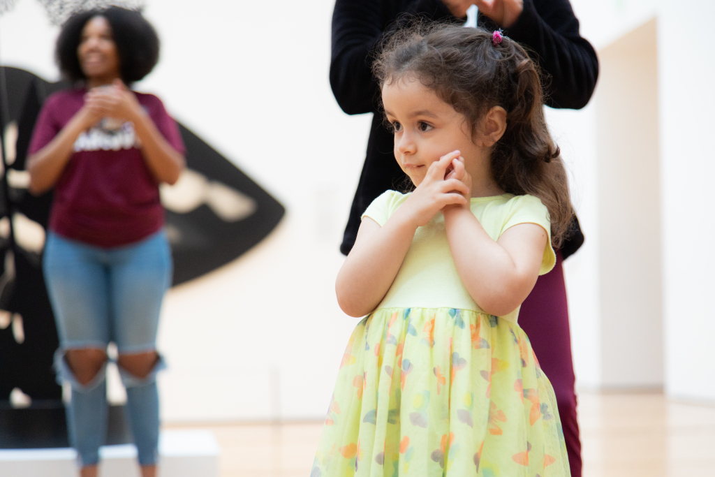 Toddler A young girl in a yellow dress stands indoors, looking to the side with her hands clasped near her face. Two adults are visible in the blurred background, one clapping and another standing behind her.