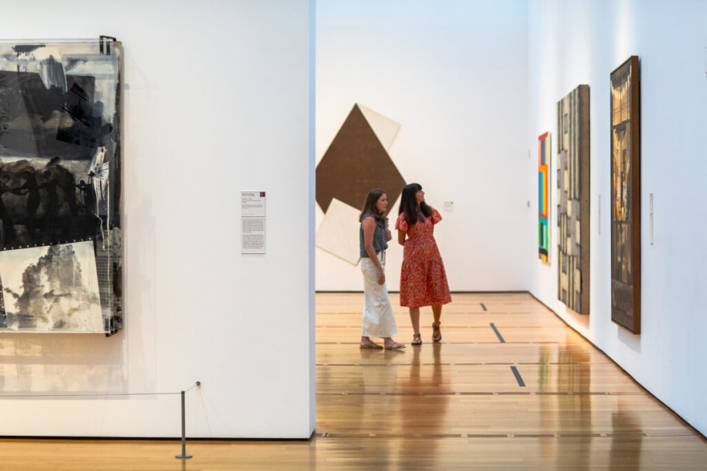 Hm Educators night august 56 O3 Two women stand and talk in a modern art gallery, surrounded by abstract paintings on white walls and a polished wooden floor. Two women stand and talk in a modern art gallery, surrounded by abstract paintings on white walls and a polished wooden floor.