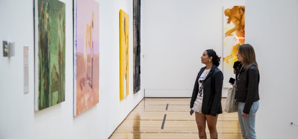 Two women stand in an art gallery, observing colorful abstract paintings displayed on white walls. The floor is wooden, and the atmosphere is quiet and contemplative.