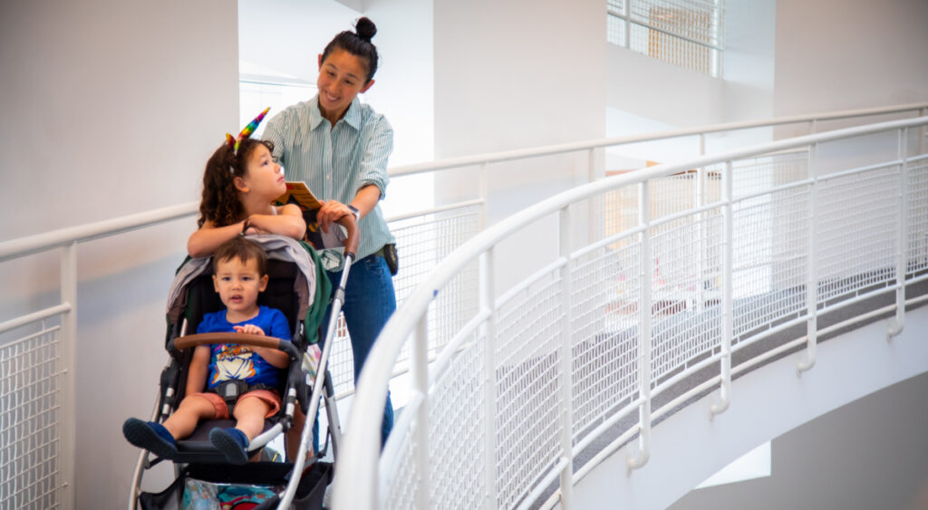 Accessforall Awhitfield June202445 O3 A woman pushes a stroller with two young children, one wearing a unicorn headband, along a curved, white indoor walkway with mesh railings.