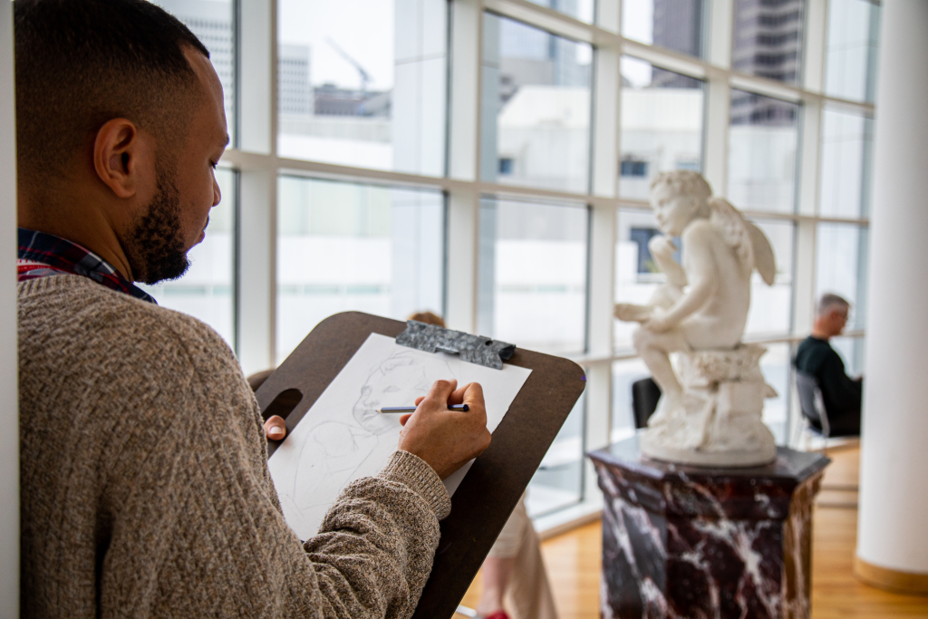 A man sketches a marble statue of a seated angel inside a bright gallery with large windows. The statue is on a pedestal, and another person sits in the background.