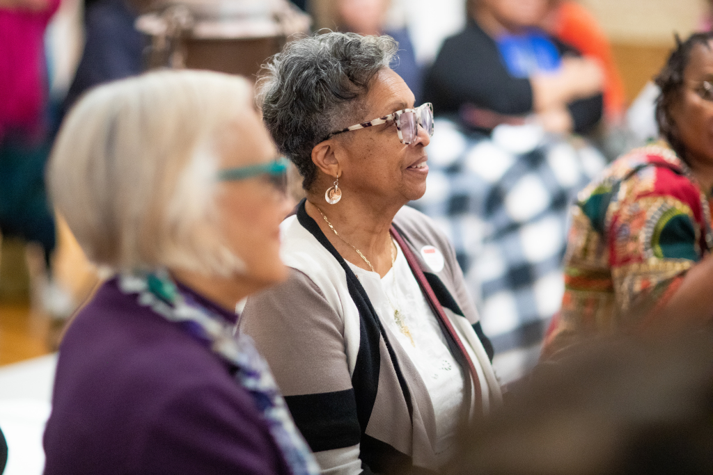 Two older women sit side by side at an indoor event, engaged and listening attentively. One wears a white and gray sweater with glasses, the other wears a purple jacket and scarf. Other people are blurred in the background.