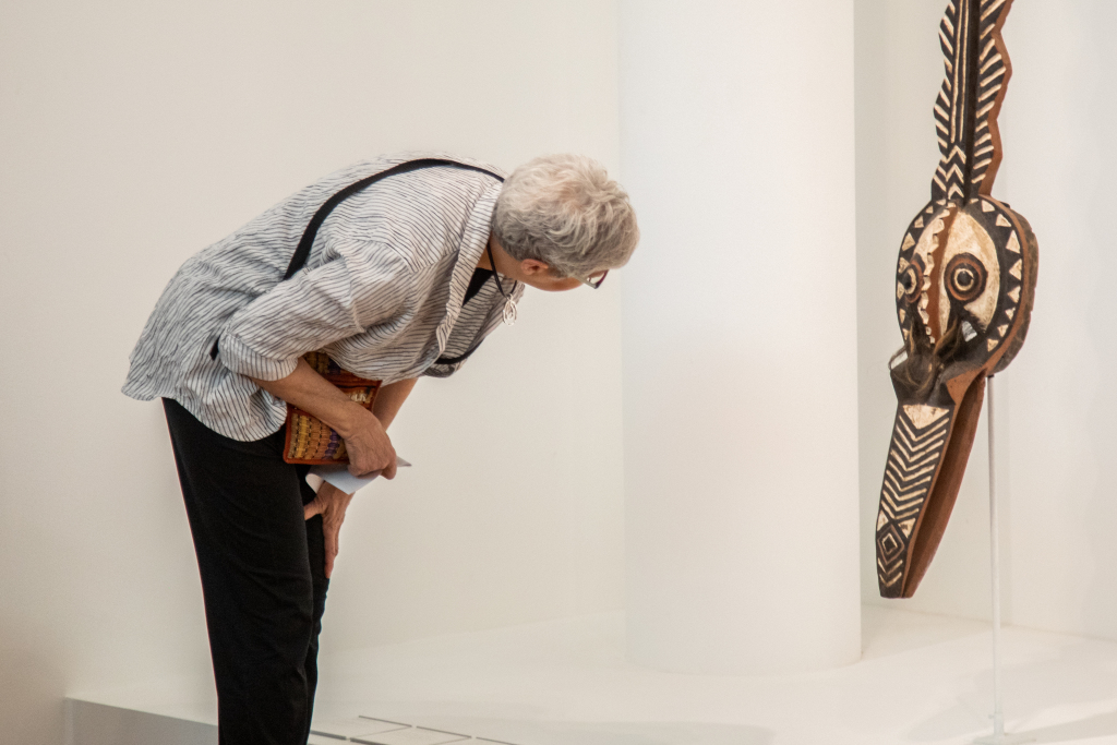 Eve Edu Lifelong Learning Celebration Lll 202348 O4 An older person with gray hair, wearing glasses and a striped shirt, bends forward to closely examine a patterned tribal mask displayed on a white wall in a gallery or museum.