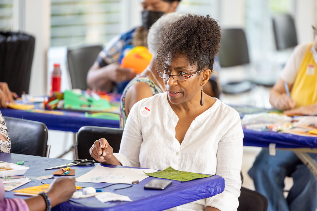 A woman wearing glasses and a white shirt sits at a table, drawing or writing on paper. She is surrounded by art supplies, with other people working on crafts in the background.