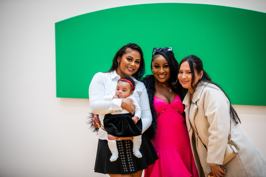 Three women pose together indoors, smiling at the camera. One woman holds a baby dressed in a red and black outfit. A large green geometric artwork hangs on the white wall behind them.