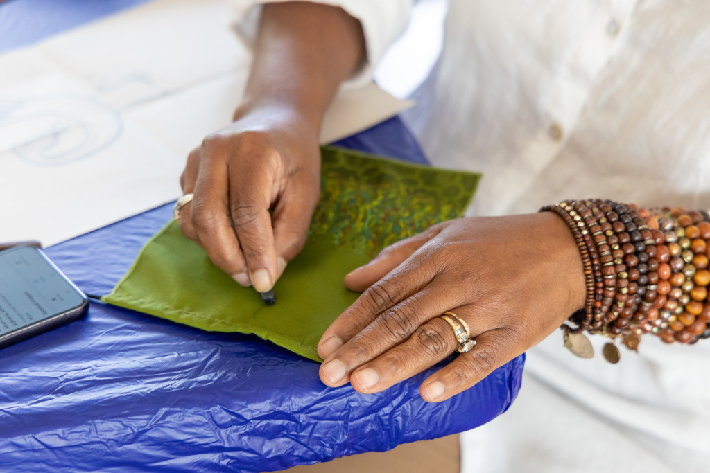 A person wearing beaded bracelets and rings draws on a green fabric square with a black pen, while sitting at a table covered with a blue tablecloth. A smartphone is visible beside their hands.