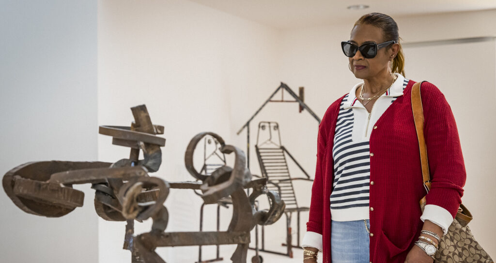 A woman wearing sunglasses, a red cardigan, and a striped shirt observes abstract metal sculptures in an art gallery. She carries a patterned bag and appears thoughtful.