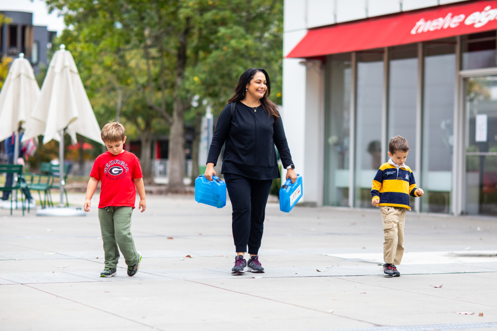 A woman walks outdoors carrying two blue lunchboxes, accompanied by two young boys, one in a red shirt and the other in a striped shirt. They are walking on a paved area near trees and a building.
