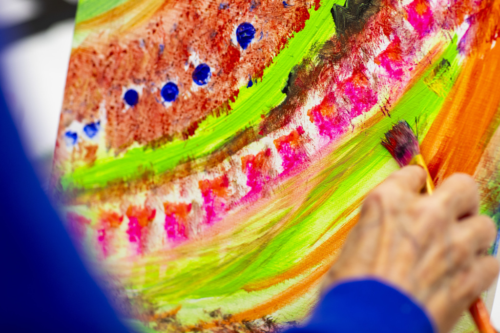 Painting Colorandtexture A close-up of a hand holding a paintbrush, painting bright, colorful abstract patterns with green, pink, orange, and blue on a canvas. The artist’s hand is slightly blurred in the foreground.