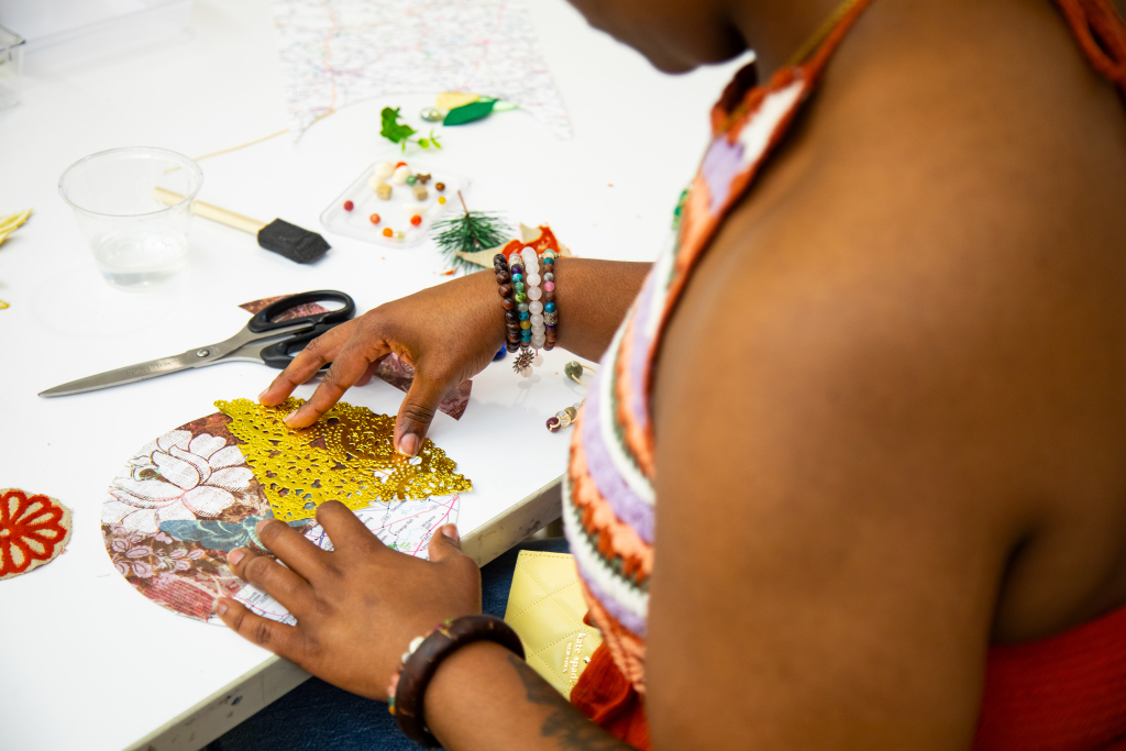 A person decorates a circular piece of paper with lace and fabric scraps at a craft table. Scissors, beads, and other art supplies are scattered nearby. The person wears a striped top and beaded bracelets.