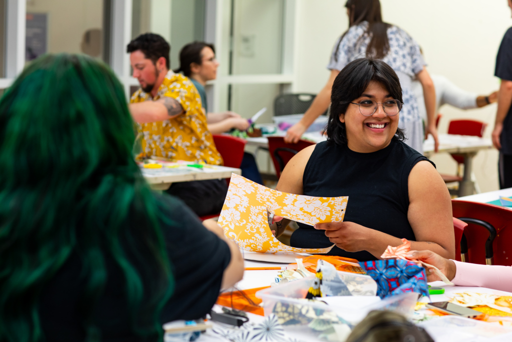 art making collage A person smiles while holding patterned paper at a table, surrounded by others engaged in creative craft activities in a brightly lit room.