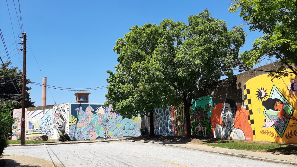 20240823 121612 Photo By John Dirga A street scene featuring colorful murals on building walls, including abstract designs and portraits, with large green trees and a clear blue sky overhead. A smokestack and tower are visible in the background.