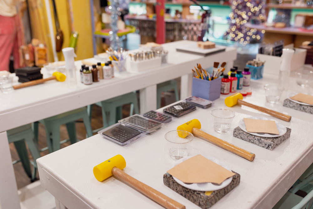 Workshop Hce 12 A brightly lit craft workshop features tables set with yellow mallets, leather pieces, small glass cups, paints, brushes, and various crafting tools, ready for a hands-on art or leatherworking activity.