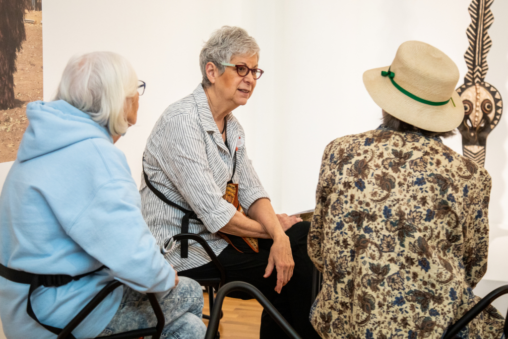 Eve Edu Lifelong Learning Celebration Lll 202328 O4 Three elderly women sit together and talk in an art gallery. One faces the camera while the others sit with their backs turned. Artwork is visible on the wall behind them.