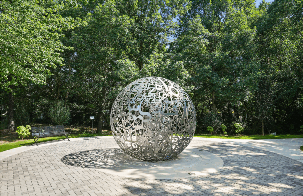 A large, spherical sculpture made of interwoven metal letters stands on a circular paved area, surrounded by trees and two benches in a park setting.