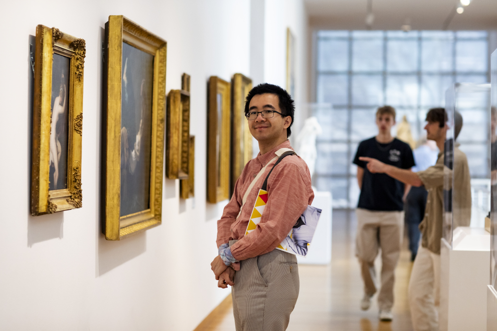 A person with glasses and a pink shirt stands smiling in an art gallery, holding a floral tote bag. Framed paintings line the wall, and two people in the background look at art and point.