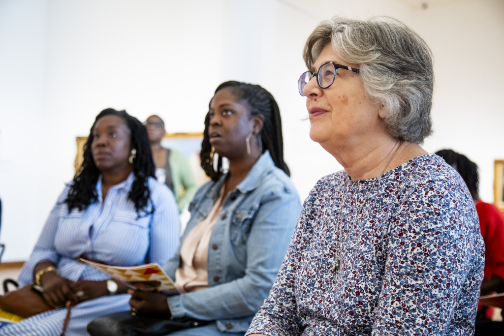 Three women sit indoors, attentively listening to something out of frame. One woman in front wears glasses and a floral top; the others behind her wear a striped dress and a denim jacket. The background is softly blurred.