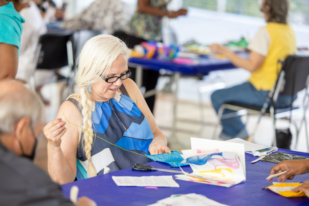 An older woman with long white hair and glasses smiles while engaging in a craft activity at a table, surrounded by colorful art supplies and other participants in a bright, communal space.