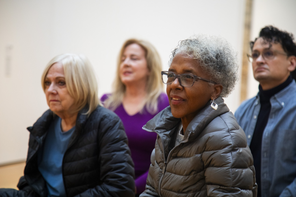 Four adults sit indoors, attentively listening. Two women with light hair are in the front and back left, a woman with short curly gray hair and glasses is in the front right, and a man with curly dark hair and glasses is in the back right.