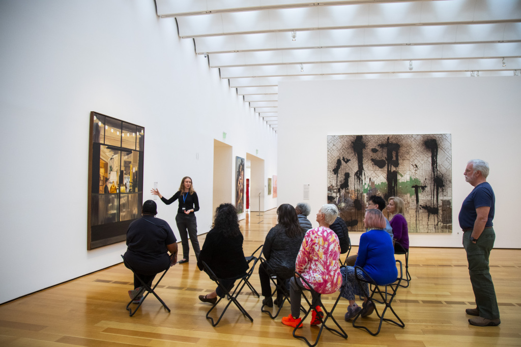 A museum guide gestures while speaking to a seated group of people in an art gallery, with large contemporary artworks displayed on white walls and natural light streaming through the ceiling.