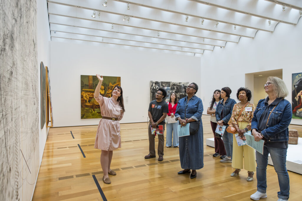 A museum guide gestures toward a large artwork on the wall while a group of attentive visitors stands and listens in a bright, modern gallery space with wooden floors and high ceilings.