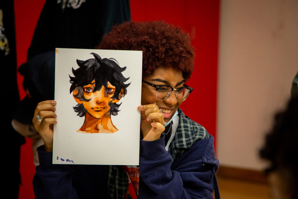 A person with curly hair and glasses smiles while holding up a drawing of an animated character’s face with expressive eyes and dark hair. Red lockers and another person are visible in the background.