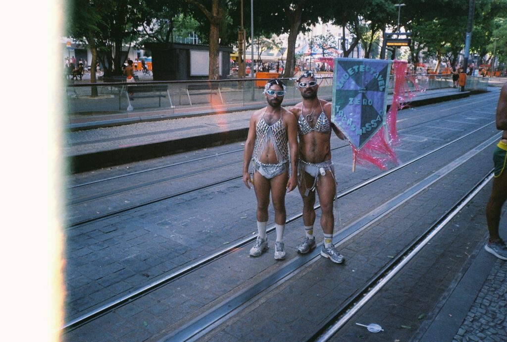 Two people in matching silver sequin outfits, sunglasses, and boots stand on tram tracks holding a colorful banner at a street festival with trees, spectators, and city elements in the background.