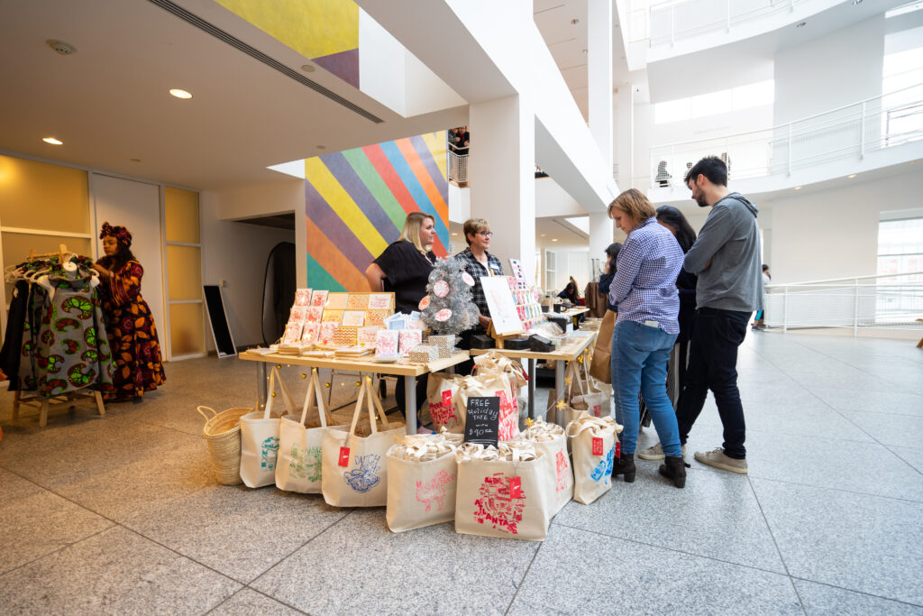 Holiday Marketin A group of people stands at a vendor table indoors, looking at printed cards, art, and tote bags for sale. The bright space has modern decor and a colorful striped wall in the background.