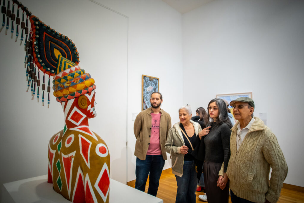 Four people stand in an art gallery, observing a colorful, patterned sculpture of a human figure with an elaborate headpiece. Paintings hang on the white walls behind them.