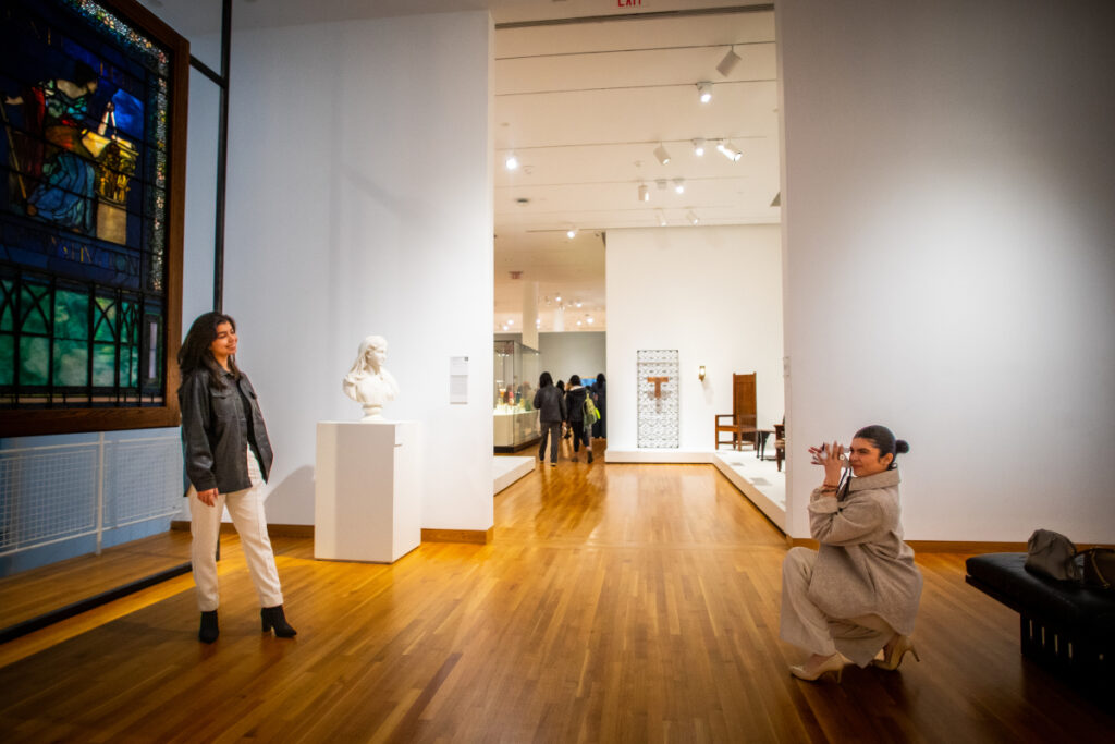 A woman poses for a photo in an art museum while another woman crouches to take her picture. Artworks, including a stained glass window and a bust, are displayed in the well-lit gallery. Other visitors are visible in the background.