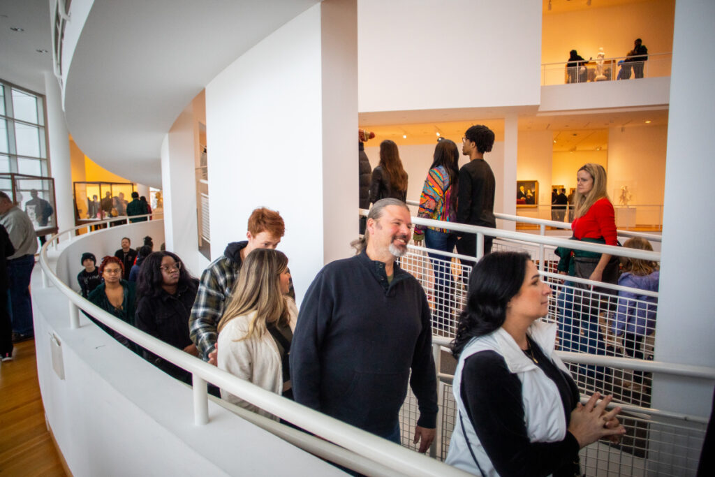 A diverse group of people walk in a line along a curved indoor ramp with white walls, inside a modern, brightly lit building with art displays and large windows.