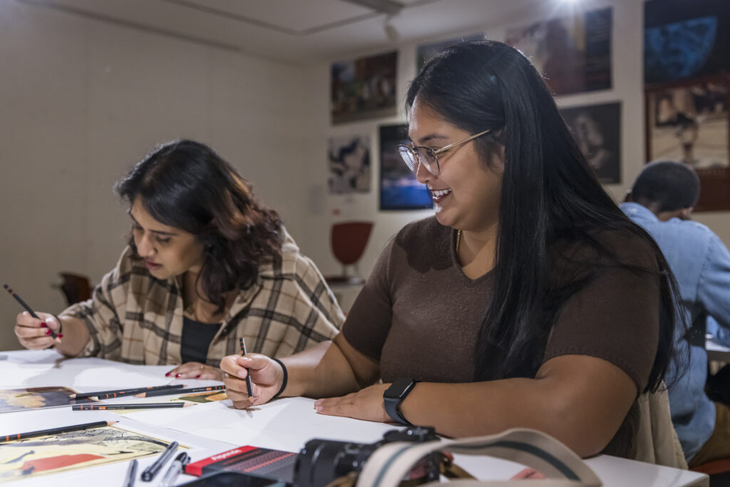 Two women sit at a table, smiling and drawing with pencils on paper. Other people are also working at desks in the background in a well-lit classroom or studio setting.