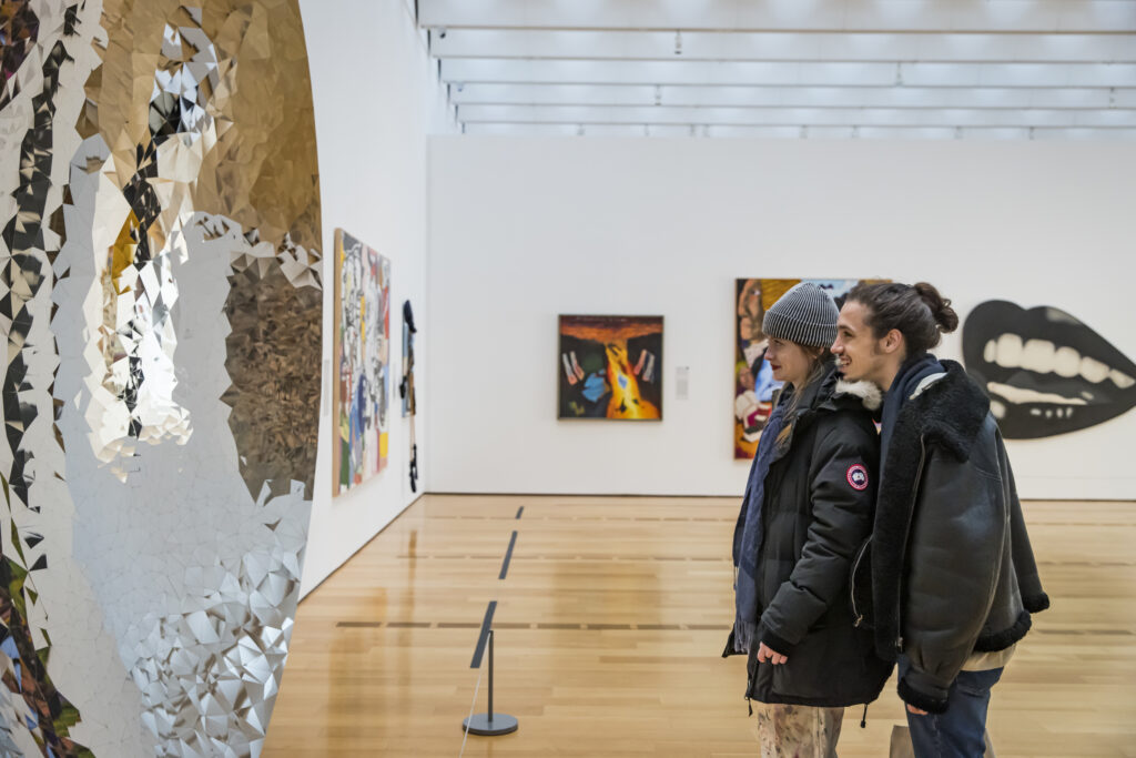 Two people stand closely together, observing a large, reflective, mosaic-like sculpture in a modern art gallery with colorful paintings on the walls and a wooden floor.