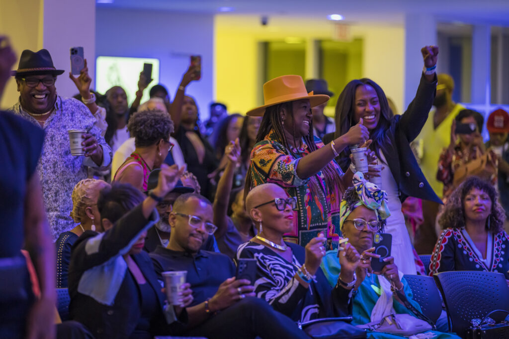A diverse group of people, some standing and some seated, cheer and raise their hands at an indoor event. Several attendees smile, hold drinks, or take photos with their phones, creating a lively and joyful atmosphere.