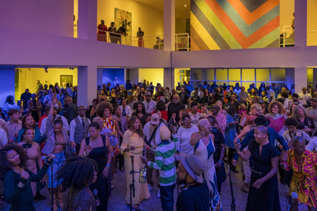 A large, diverse crowd of people enjoy a lively indoor event with colorful lighting and bold wall art. Some are dancing, singing, or talking, creating an energetic and festive atmosphere.