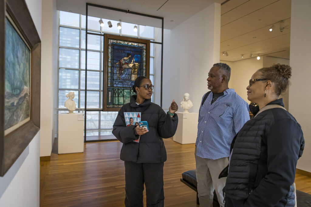 Three people stand together in an art gallery, looking at a painting on the wall. Behind them are sculptures and a large stained-glass window. They appear to be in conversation and engaged with the artwork.