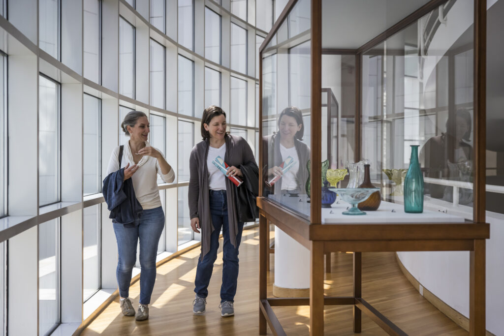 Two women walk through a modern museum gallery, smiling and talking while viewing glass art pieces displayed in a wooden and glass case next to large, curved windows.