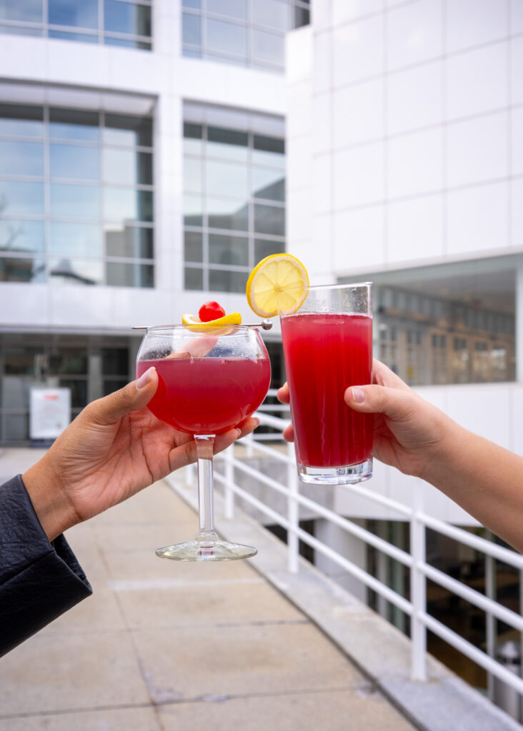 Two hands clinking glasses of bright red drinks garnished with lemon slices and a cherry, set outdoors against a modern white building with large windows.