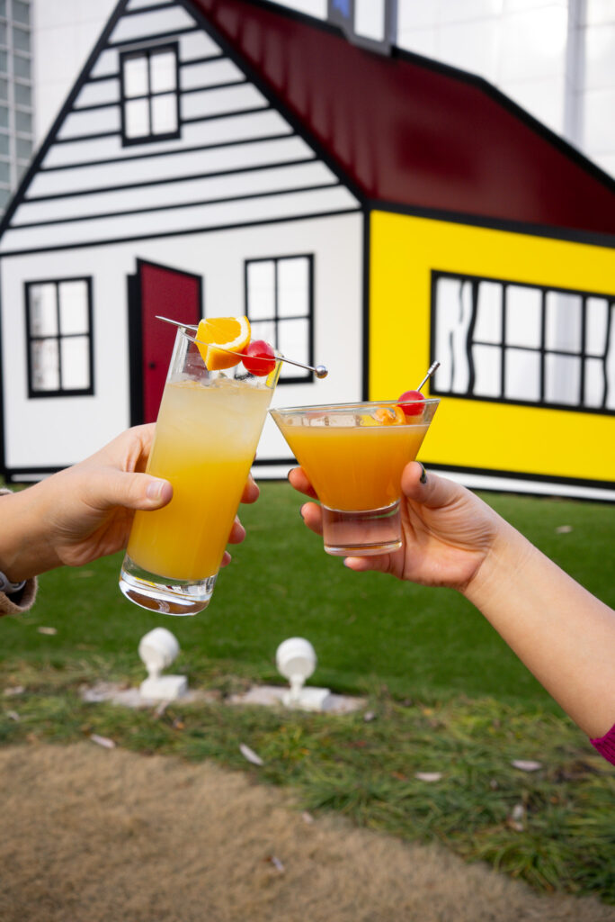 Two hands holding colorful cocktails with fruit garnishes, raised in a cheers gesture, in front of a playful, cartoon-like house with yellow, white, and red colors in the background.