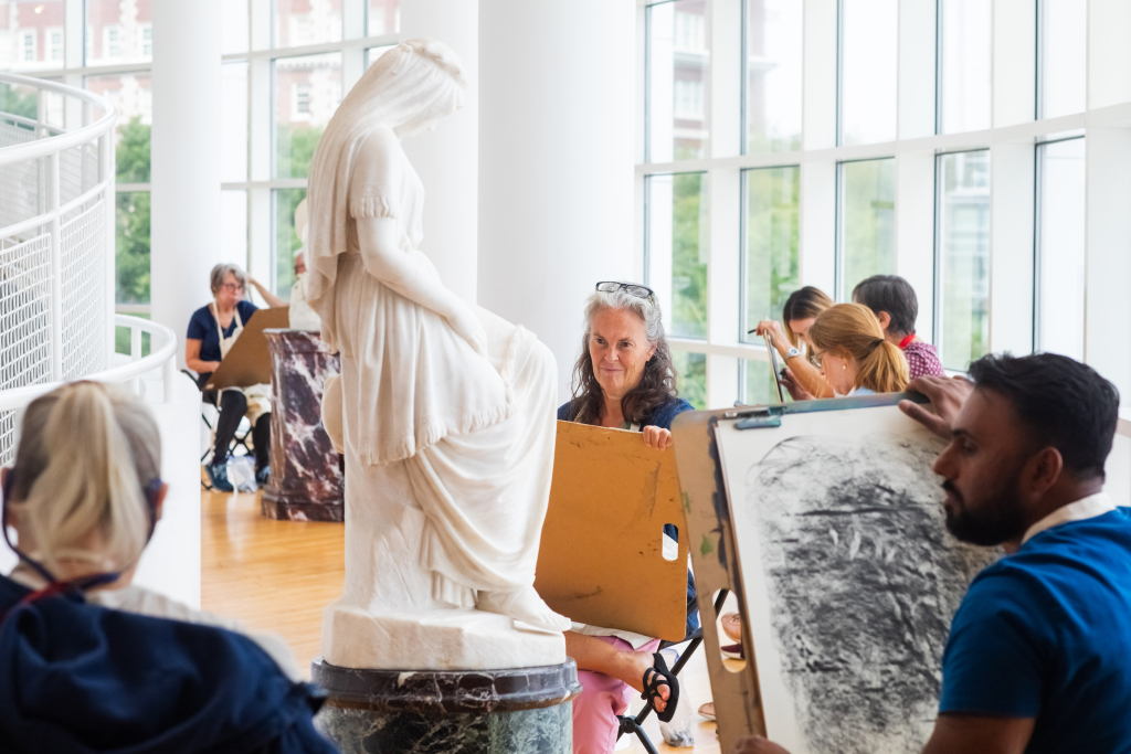 People sit on stools in a bright room sketching a white marble statue of a woman in a flowing dress. Large windows line the wall, and the artists focus on their drawings with sketchpads in hand.