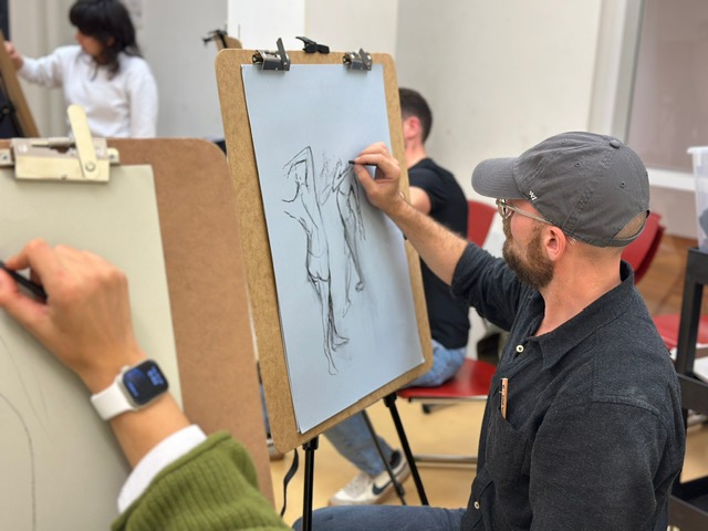 Several people sit at easels sketching a live model in a classroom or studio. A man in a gray cap draws figures on a large sheet, while another person’s hand with a smartwatch sketches nearby.