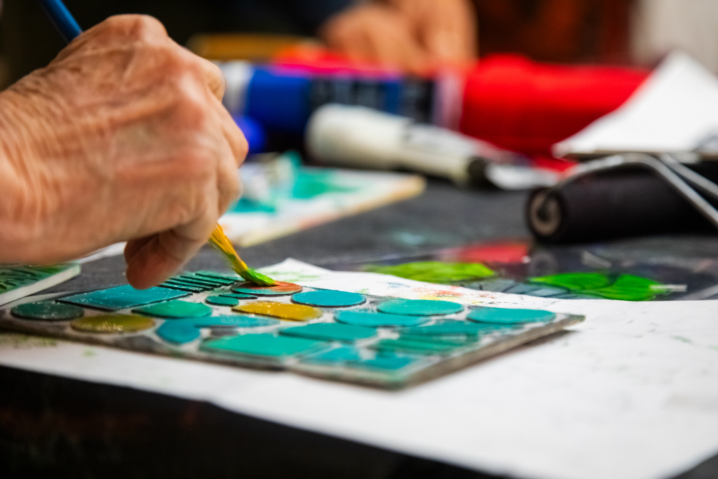 A close-up of a hand painting colorful shapes on a ceramic tile with a brush. Art supplies and bright paint tubes are visible in the blurred background.
