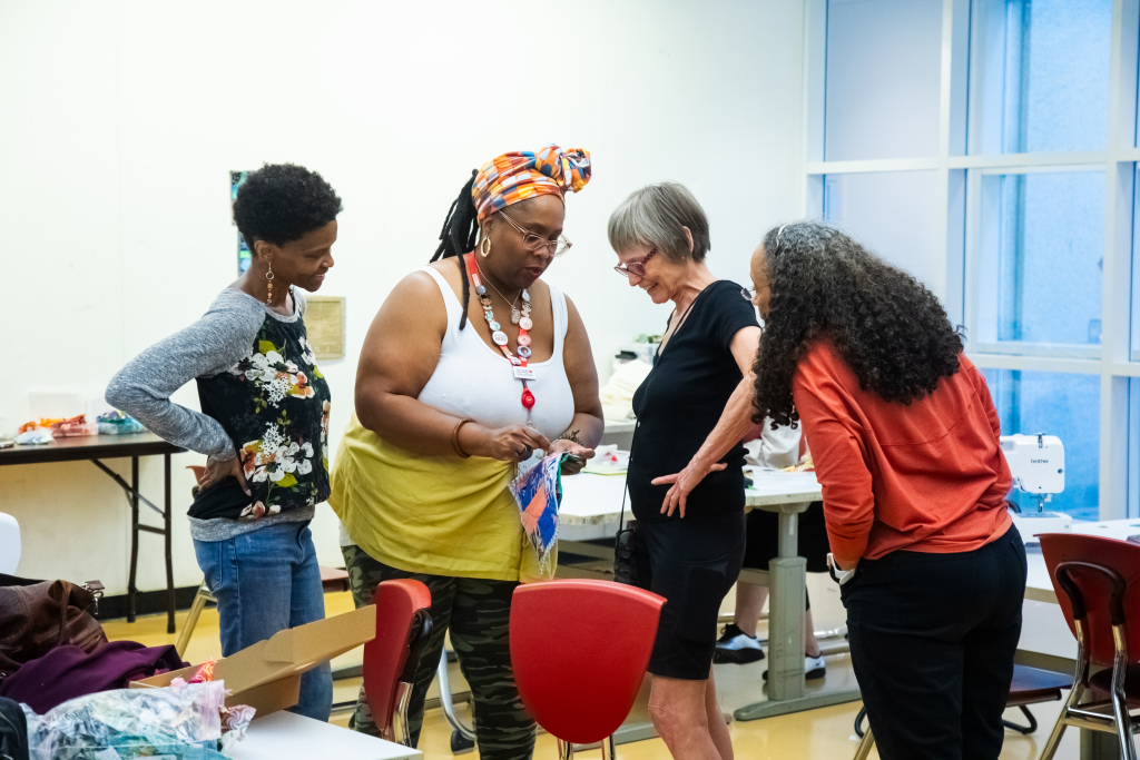 Four women stand together in a brightly lit room, engaged in conversation. One holds colorful fabric, while the others look on with interest. The setting appears to be a creative or educational workshop.