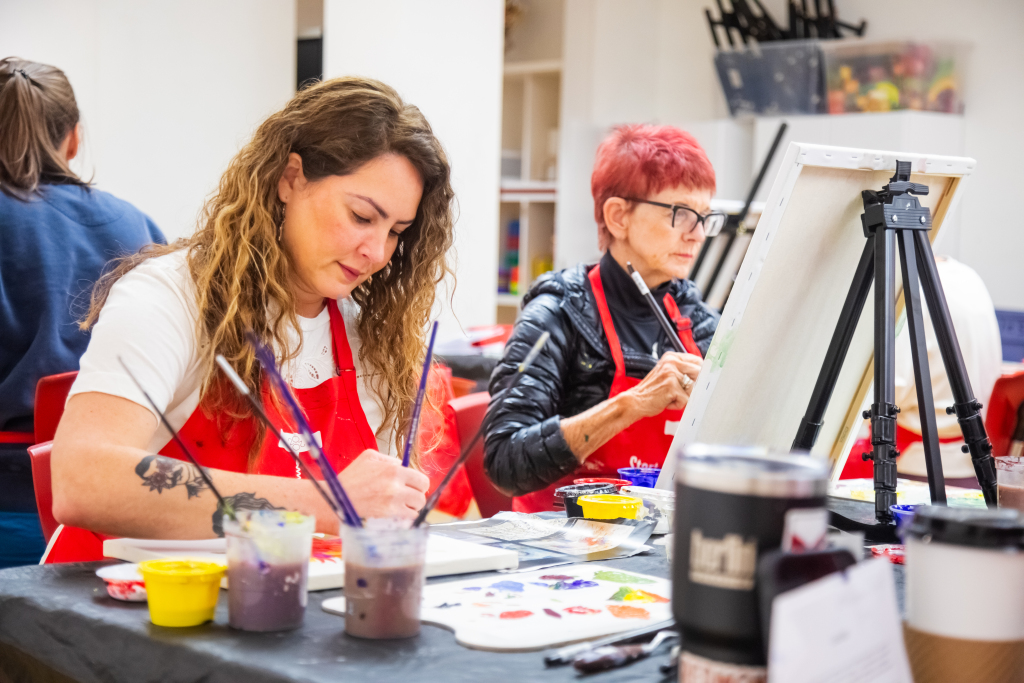 Two women sit at a table painting on canvases, focused on their artwork. They wear red aprons and are surrounded by paint supplies, brushes, and cups in a bright, indoor art studio.